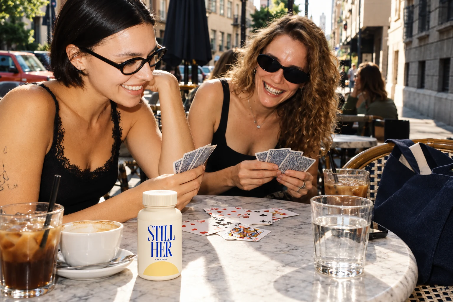 Friends laughing over cards at a café, with a STILL HER bottle beside their coffee
