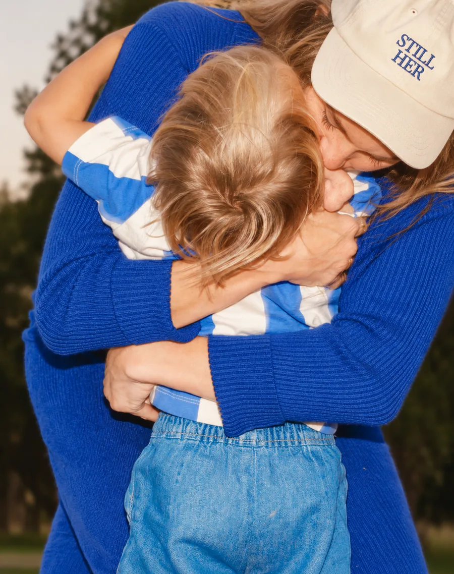 Mother in a cobalt sweater and STILL HER cap hugging her child